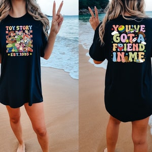 May include: Black t-shirt with a colorful Toy Story graphic on the front and the text "You've Got a Friend in Me" on the back. The shirt is worn by a person standing on a beach.