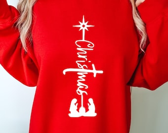 May include: A red Christmas sweatshirt with a white vertical design. The design includes a star, the word "Christmas" in script, and a nativity scene. The sweatshirt has long sleeves and a crew neck.