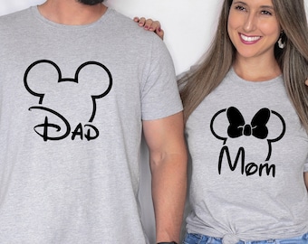 May include: Two gray t-shirts with black graphics. One shirt features a Mickey Mouse head outline with the word "DAD" below. The other shirt has a Minnie Mouse head outline with a bow and the word "Mom" below. Both shirts are short-sleeved.