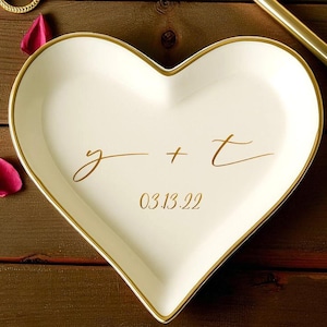 May include: A heart-shaped, white ceramic ring dish with a gold rim and gold lettering. The dish has the initials "g + t" and the date "03.13.22". Two gold rings, a gold pen, and rose petals are on a wooden surface.