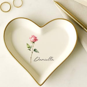 May include: Heart-shaped ceramic trinket dish with a gold rim, featuring a pink rose illustration and the name "Daniella" in script. Two gold rings are visible. A gold pen and a small box are also in the image.
