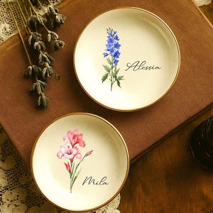 K&ouml;nnte beinhalten: Zwei kleine, runde, cremefarbene Zierteller mit goldenen R&auml;ndern. Einer zeigt eine blaue Blume und den Namen "Alessia", der andere eine rosa Blume und den Namen "Mila". Die Teller liegen auf einer braunen Oberfl&auml;che.
