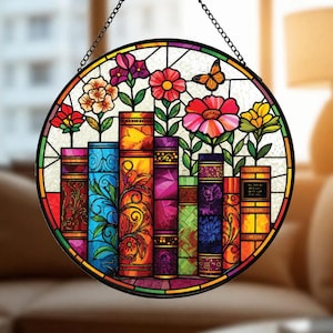 May include: A circular stained glass suncatcher featuring a row of colorful books with flowers growing from their tops and a small butterfly. The piece is framed in black metal and hangs from a thin black chain, displaying vibrant hues of red, blue, yellow, and purple.
