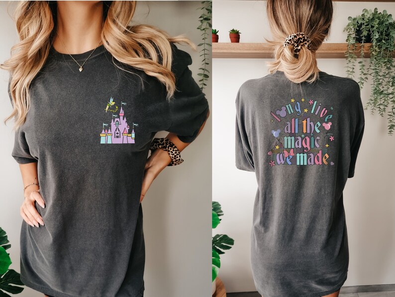 May include: Dark gray t-shirt featuring a colorful castle graphic with Tinkerbell. The back of the shirt displays the text "I long live all the magic we made" in a whimsical font with Mickey Mouse ears and stars.