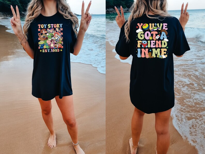 May include: Black t-shirt with a colorful Toy Story graphic on the front and the text "You've Got a Friend in Me" on the back. The shirt is worn by a person standing on a beach.