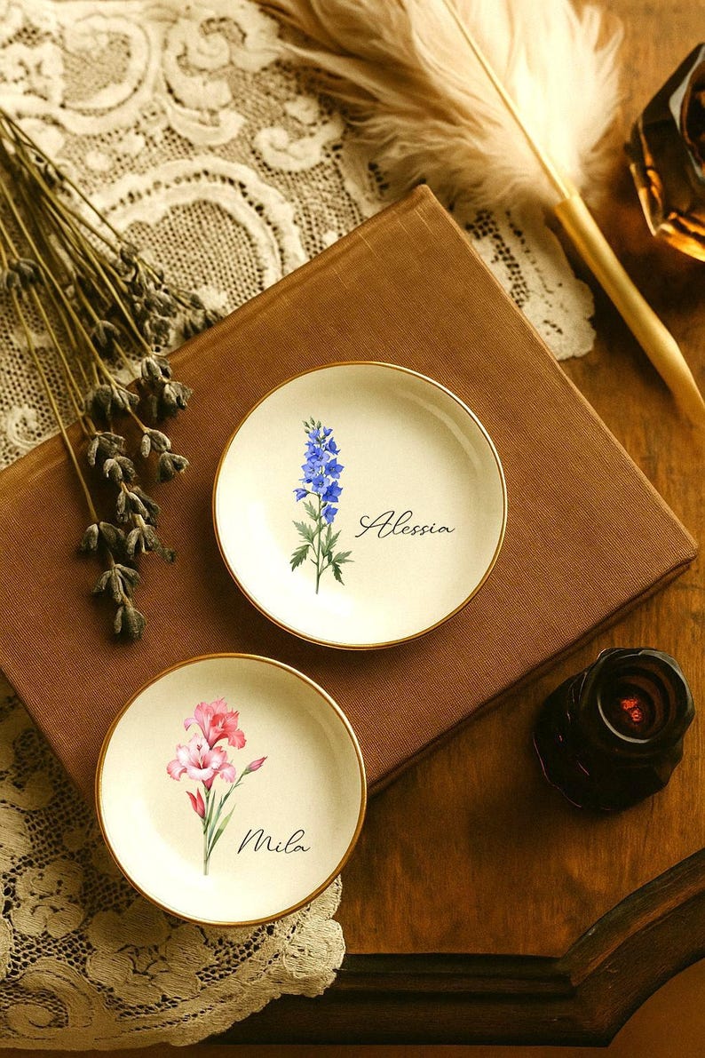 K&ouml;nnte beinhalten: Zwei kleine, runde, cremefarbene Zierteller mit goldenen R&auml;ndern. Einer zeigt eine blaue Blume und den Namen "Alessia", der andere eine rosa Blume und den Namen "Mila". Die Teller liegen auf einer braunen Oberfl&auml;che.