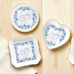 May include: White ceramic trinket dishes in round, heart, and square shapes, each with a gold rim. The dishes feature a blue floral design with two intertwined hearts and text. The text includes dates such as Jun.29.27 and Mar.23.26.