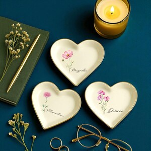 K&ouml;nnte beinhalten: Drei herzf&ouml;rmige Schmuckschalen mit Blumendesigns und goldenen R&auml;ndern. Jede Schale zeigt eine andere rosa Blume und einen Namen: Magnolia, Clementine und Desiree. Eine brennende Kerze, ein Stift und eine Brille sind ebenfalls zu sehen.
