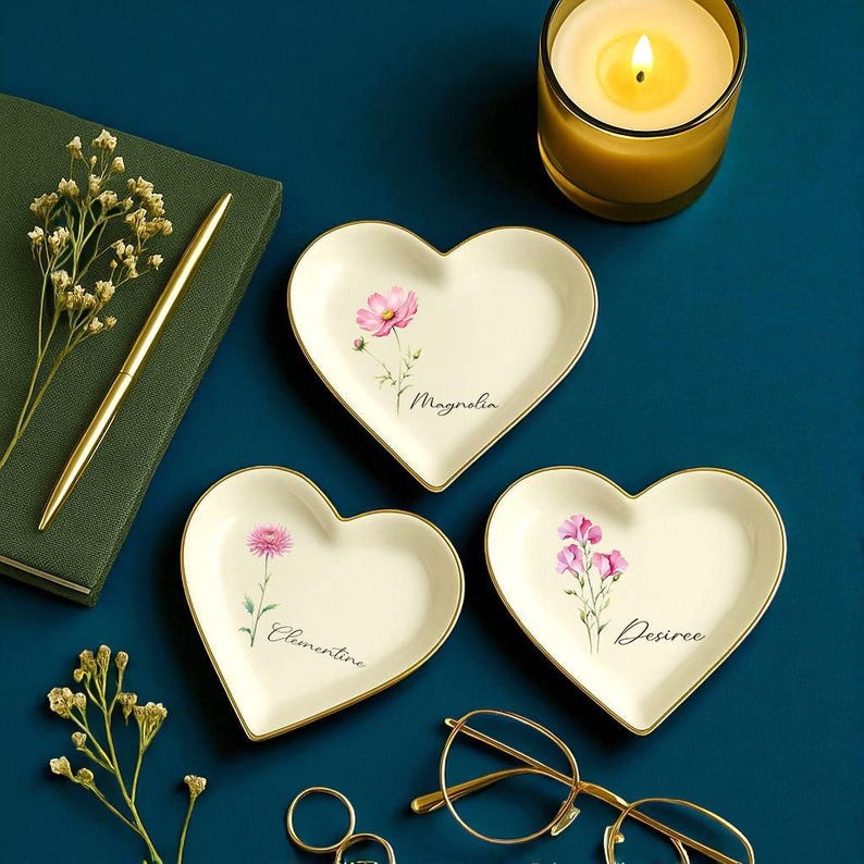 K&ouml;nnte beinhalten: Drei herzf&ouml;rmige Schmuckschalen mit Blumendesigns und goldenen R&auml;ndern. Jede Schale zeigt eine andere rosa Blume und einen Namen: Magnolia, Clementine und Desiree. Eine brennende Kerze, ein Stift und eine Brille sind ebenfalls zu sehen.