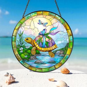 May include: Round stained glass window art featuring a colorful turtle with a purple and green shell, a blue bow, and a butterfly. The artwork depicts a serene scene with water, rocks, and plants, all set against a sunny sky. The window is framed with a chain for hanging.