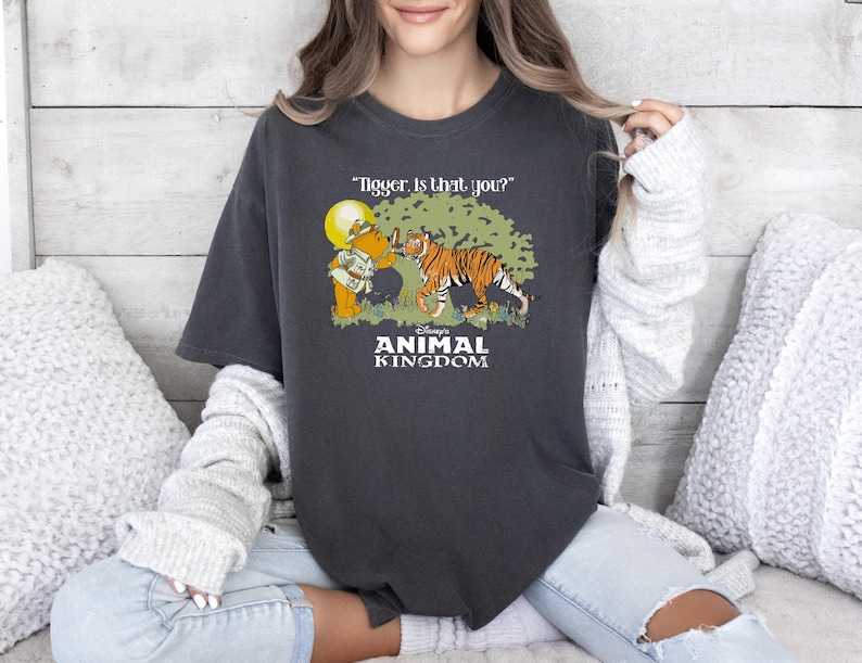May include: A dark grey t-shirt with a vintage-style graphic of Winnie the Pooh and Tigger in front of a tree. The text "Tigger, is that you?" is above the image and "Disney's Animal Kingdom" is below the image.