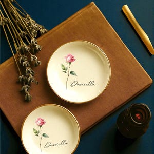 May include: Two small, off-white ceramic plates with gold rims, each featuring a pink rose illustration and the name "Daniella" in script. The plates rest on a brown surface, with a dark blue background. A gold-colored item and a dark brown object are also present.
