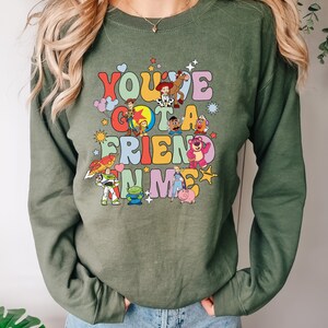 May include: Olive green sweatshirt featuring the phrase "You've Got A Friend In Me" with colorful cartoon characters. The design includes characters like Woody, Buzz Lightyear, and others from a popular animated movie.
