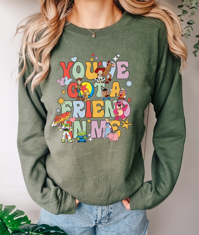 May include: Olive green sweatshirt featuring the phrase "You've Got A Friend In Me" with colorful cartoon characters. The design includes characters like Woody, Buzz Lightyear, and others from a popular animated movie.