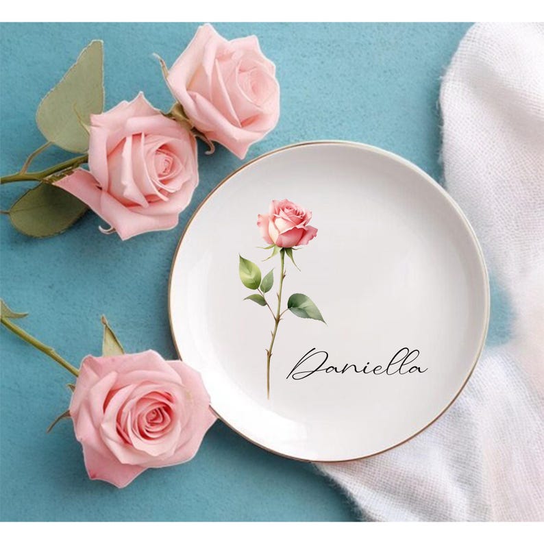 May include: A white ceramic plate with a gold rim, featuring a watercolor rose illustration and the name "Daniella" in script. Pink roses and green leaves are arranged around the plate on a blue surface.