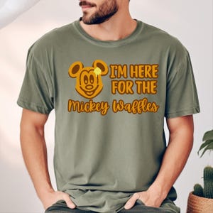 May include: Olive green t-shirt with a waffle design featuring Mickey Mouse and the text "I'm Here For The Mickey Waffles" in brown. The shirt is a comfortable, casual style.