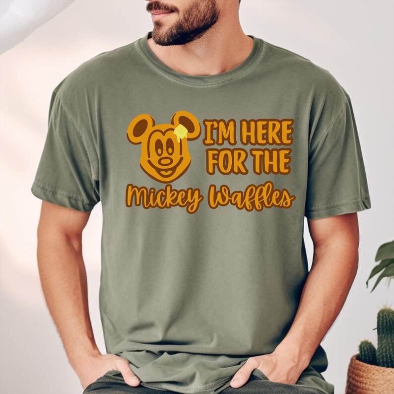 May include: Olive green t-shirt with a waffle design featuring Mickey Mouse and the text "I'm Here For The Mickey Waffles" in brown. The shirt is a comfortable, casual style.