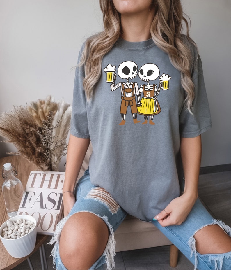 May include: Gray t-shirt with a graphic of two skeletons dressed in traditional German Oktoberfest attire, holding beer steins.