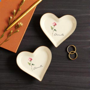 May include: Two heart-shaped trinket dishes with a pink rose design and gold trim. Each dish has the name "Daniella" written on it. A gold pen and a small notebook are in the background, along with two gold rings.