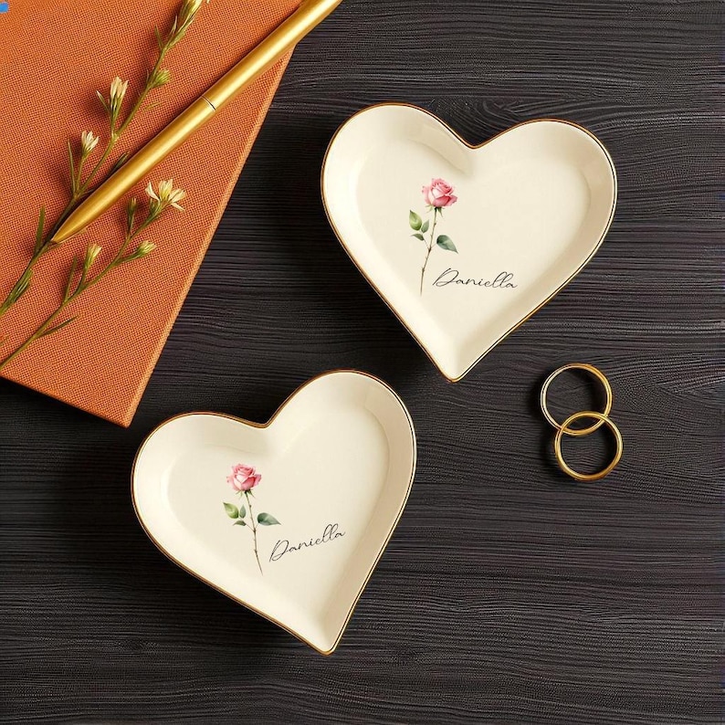 May include: Two heart-shaped trinket dishes with a pink rose design and gold trim. Each dish has the name "Daniella" written on it. A gold pen and a small notebook are in the background, along with two gold rings.
