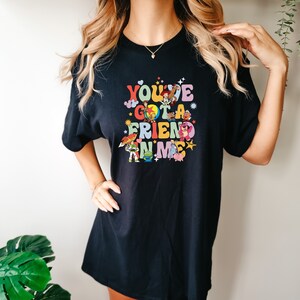 May include: Black t-shirt featuring the phrase "You've Got a Friend in Me" in colorful, retro-style lettering. The design includes cartoon characters from the Toy Story movie. A fun and playful graphic tee.