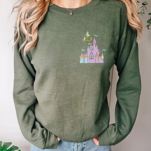 May include: Olive green sweatshirt featuring a colorful graphic of a fairytale castle with Tinkerbell. The castle has pink, purple, blue, and yellow accents. The sweatshirt has long sleeves and a crew neck.