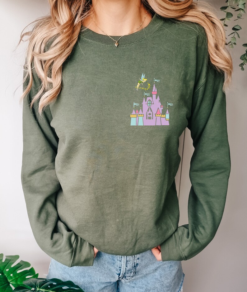 May include: Olive green sweatshirt featuring a colorful graphic of a fairytale castle with Tinkerbell. The castle has pink, purple, blue, and yellow accents. The sweatshirt has long sleeves and a crew neck.
