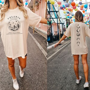 May include: A person wearing a light beige t-shirt with a black graphic print on the front that says "Velaris City of Stars" and a black graphic print on the back with a series of moons and stars. The person is walking down a street with colorful lanterns hanging overhead.