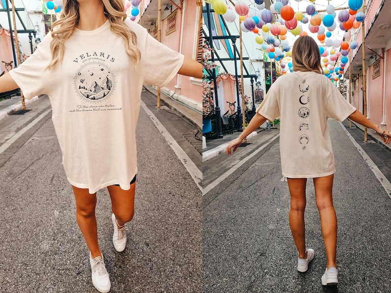 May include: A person wearing a light beige t-shirt with a black graphic print on the front that says "Velaris City of Stars" and a black graphic print on the back with a series of moons and stars. The person is walking down a street with colorful lanterns hanging overhead.