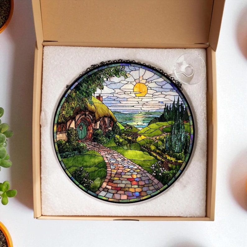 May include: Circular stained glass artwork showing a hobbit hole scene with a colourful stone path leading to a green door. The piece features a vibrant landscape with a yellow sun, blue sky, and rolling green hills. It is set in a black metal frame.