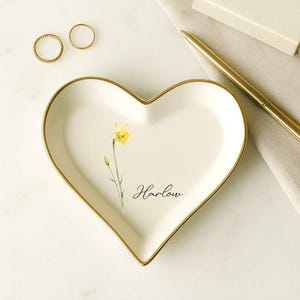 K&ouml;nnte beinhalten: Herzf&ouml;rmige Schmuckschale mit goldenem Rand, mit einem Aquarell-Osterglockenmotiv und dem Schriftzug "Harlow". Zwei goldene Ringe sind sichtbar. Ein goldener Stift und eine kleine Schachtel sind ebenfalls vorhanden.