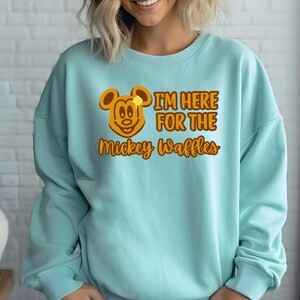 May include: A light blue sweatshirt with the text "I'M HERE FOR THE Mickey Waffles" in orange. The design includes a cartoon Mickey Mouse head with a waffle and butter. The sweatshirt is a casual style.