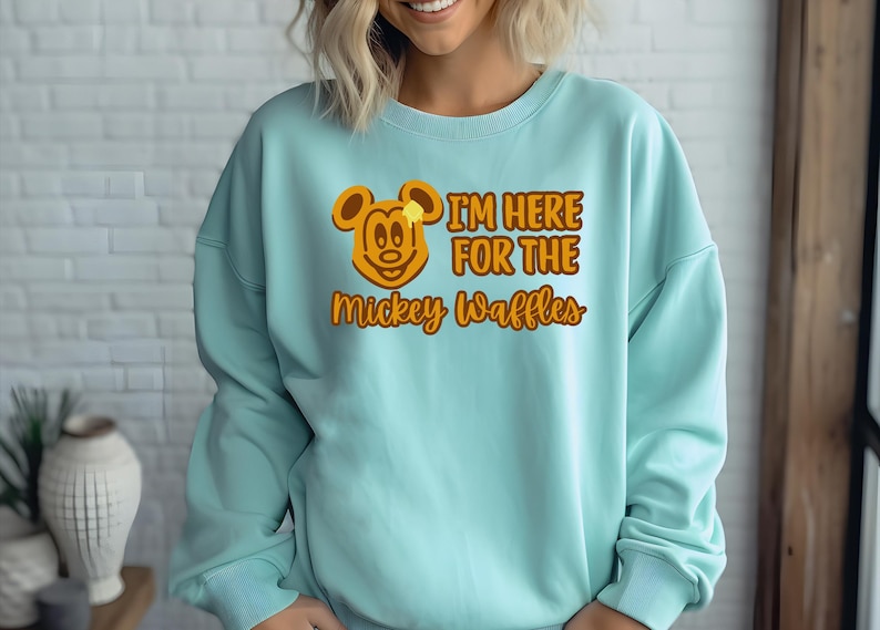May include: A light blue sweatshirt with the text "I'M HERE FOR THE Mickey Waffles" in orange. The design includes a cartoon Mickey Mouse head with a waffle and butter. The sweatshirt is a casual style.