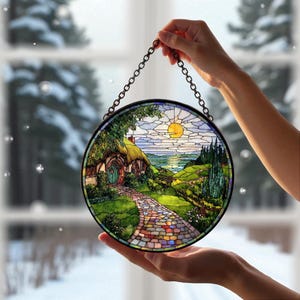 May include: Circular stained glass window art showing a hobbit house with a thatched roof, a colourful stone path, and a sunny landscape. The artwork is framed with a black metal chain for hanging. The window art is held up by a person's hands.