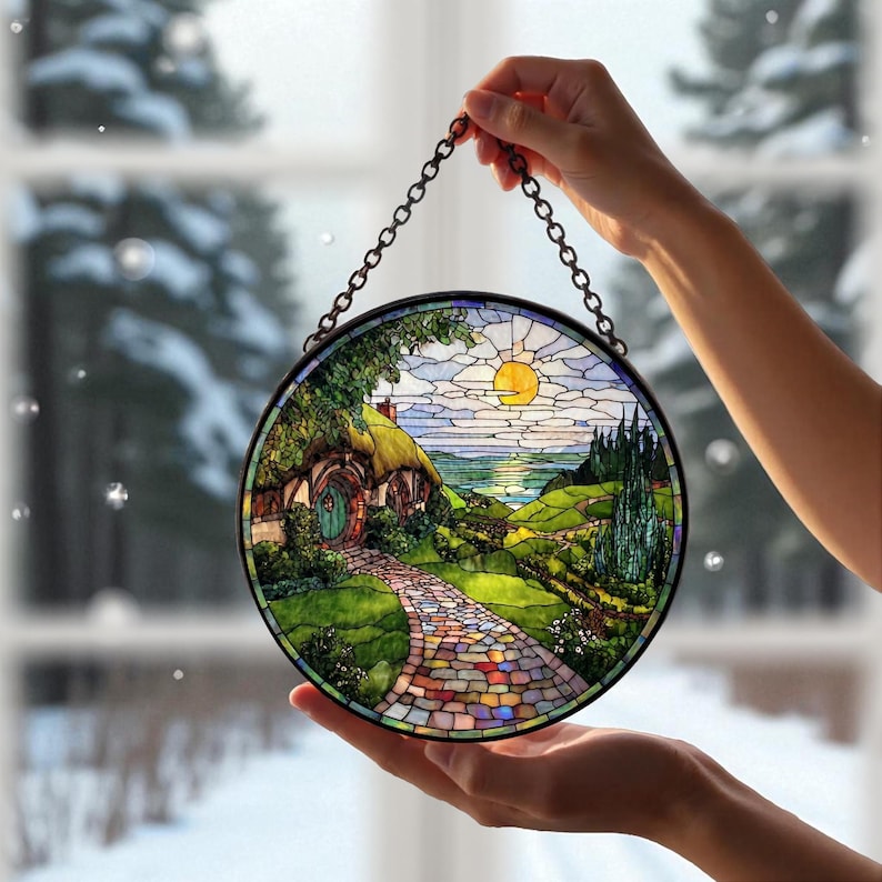 May include: Circular stained glass window art showing a hobbit house with a thatched roof, a colourful stone path, and a sunny landscape. The artwork is framed with a black metal chain for hanging. The window art is held up by a person's hands.