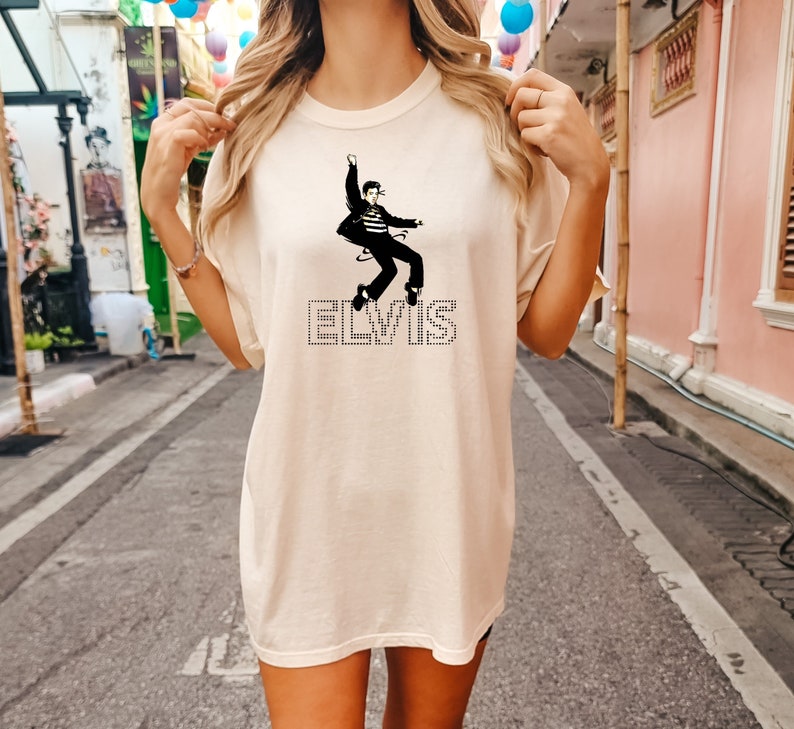 May include: A cream coloured t-shirt featuring a black silhouette of Elvis Presley in a dancing pose. The word "ELVIS" is spelled out in black rhinestones below the image.