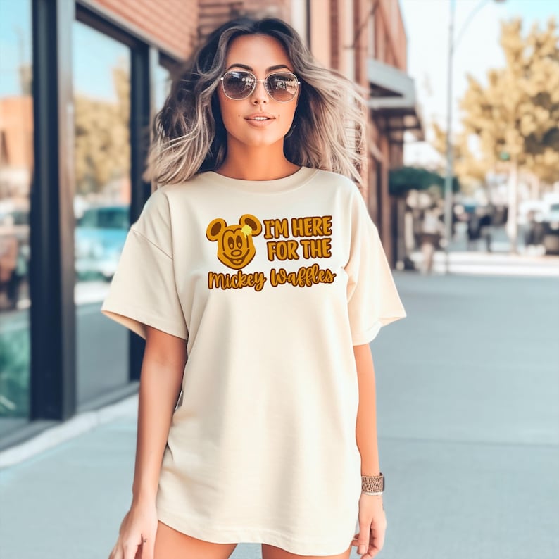 May include: Cream-colored t-shirt featuring a Mickey Mouse waffle design with the text "I'M HERE FOR THE Mickey Waffles" in brown and yellow. The shirt has short sleeves and a relaxed fit.