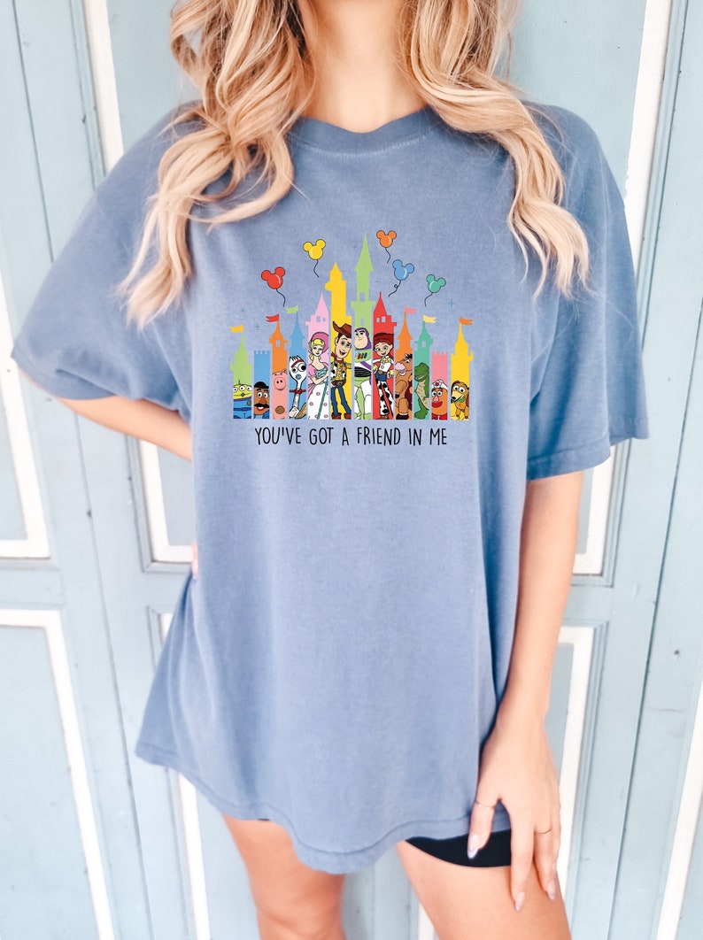 May include: A light blue t-shirt with a graphic of the Toy Story characters in front of a colorful castle silhouette. The text "You've got a friend in me" is printed below the graphic.