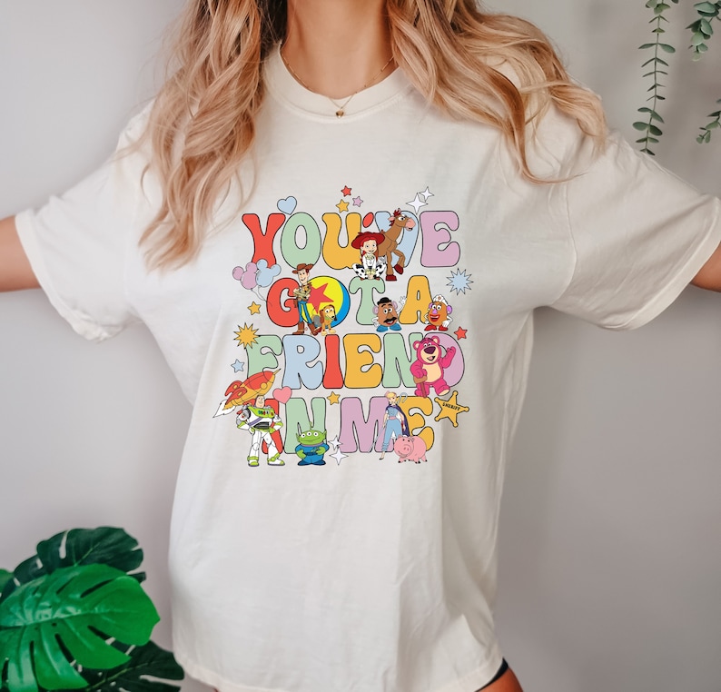 May include: Cream-colored t-shirt featuring a colorful graphic of characters from the movie "Toy Story." The text "You've Got a Friend in Me" is displayed in a retro font. The design includes Woody, Buzz Lightyear, and other characters.