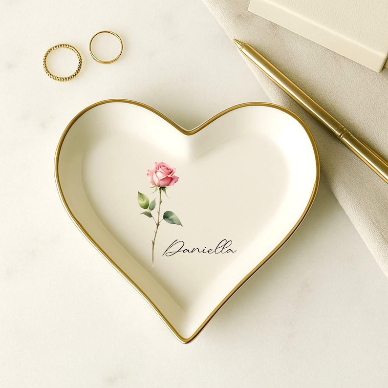 May include: Heart-shaped ceramic trinket dish with a gold rim, featuring a pink rose illustration and the name "Daniella" in script. Two gold rings are visible. A gold pen and a small box are also in the image.