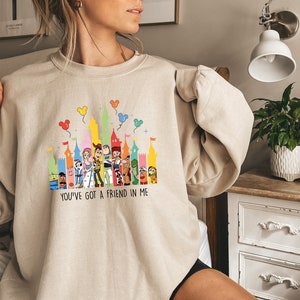 May include: A beige sweatshirt with a colorful graphic of the Disney castle and characters from Toy Story. The text "You've got a friend in me" is printed below the graphic.