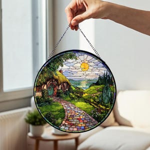 May include: Circular stained glass panel showing a hobbit hole scene with a path leading to a green door. The artwork features a sunny sky, rolling hills, and a colourful stone path, all framed in a black metal circle.