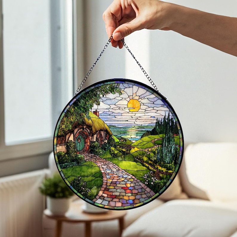 May include: Circular stained glass panel showing a hobbit hole scene with a path leading to a green door. The artwork features a sunny sky, rolling hills, and a colourful stone path, all framed in a black metal circle.