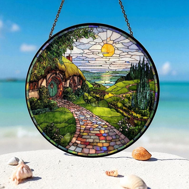 May include: Circular stained glass window showing a hobbit house with a thatched roof, a stone path, and a sunny landscape. The artwork is framed in black and suspended by a chain. The scene includes a bright yellow sun and a blue sky.