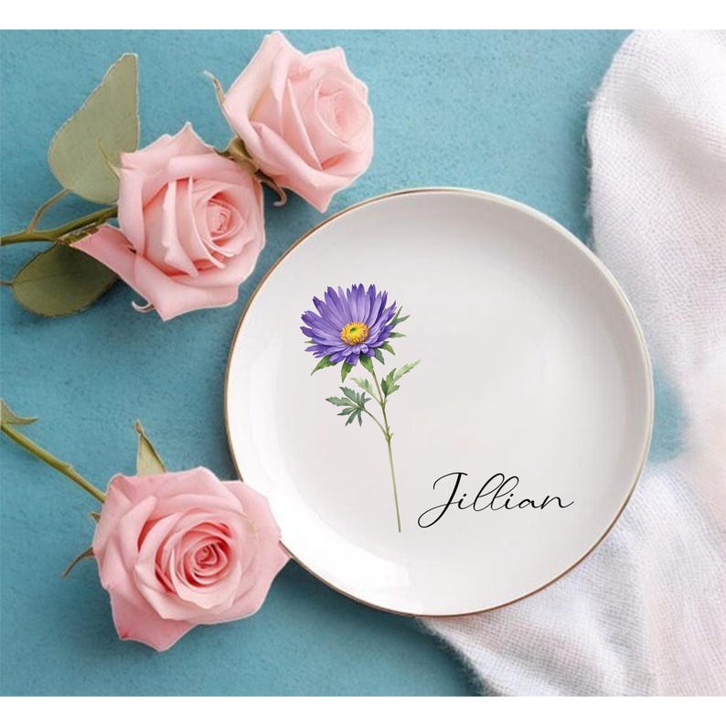 K&ouml;nnte beinhalten: Wei&szlig;e Keramikplatte mit goldenem Rand, mit einer lila Blumenillustration und dem Namen "Jillian" in Schreibschrift. Rosa Rosen und gr&uuml;ne Bl&auml;tter sind um die Platte auf einer blauen Oberfl&auml;che angeordnet.