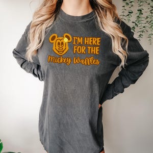May include: Dark gray long-sleeve shirt with a graphic of Mickey Mouse and the text "I'M HERE FOR THE Mickey Waffles" in orange. The shirt has a vintage, washed look and a relaxed fit.