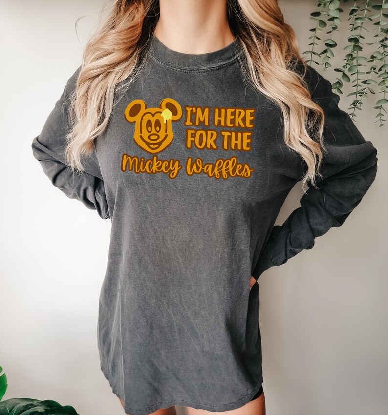 May include: Dark gray long-sleeve shirt with a graphic of Mickey Mouse and the text "I'M HERE FOR THE Mickey Waffles" in orange. The shirt has a vintage, washed look and a relaxed fit.