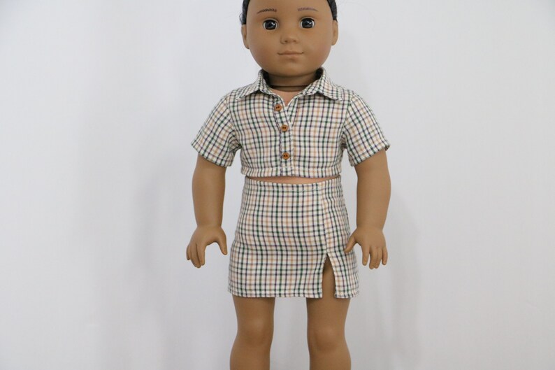 May include: A doll wearing a plaid shirt and skirt set. The shirt is a cropped style with short sleeves and a button closure. The skirt is a mini skirt with a side slit.