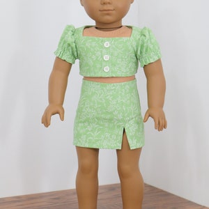 May include: A green floral print crop top and skirt set for a doll. The top has a square neckline and short puff sleeves. The skirt has a side slit.