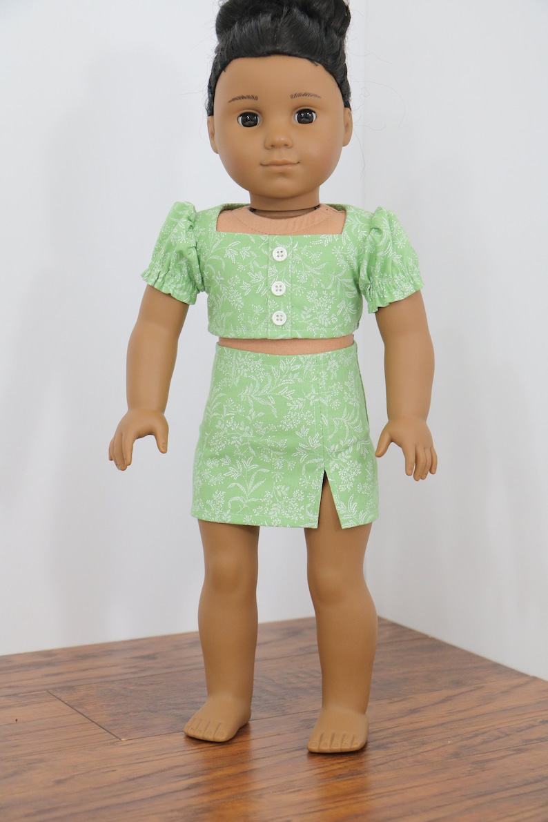 May include: A green floral print crop top and skirt set for a doll. The top has a square neckline and short puff sleeves. The skirt has a side slit.